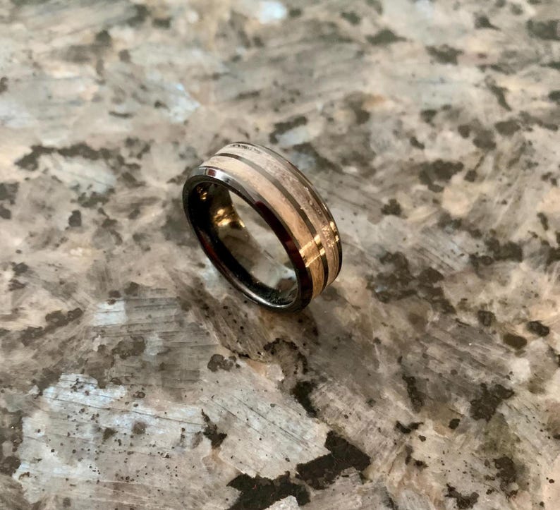 May include: A black ring with a wooden inlay and a silver band. The ring has a unique design with alternating light and dark wood grain patterns. The ring is likely made of tungsten carbide and wood.