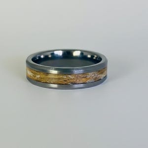May include: A dark gray ring with a light-colored wood-like inlay. The ring has a smooth, polished finish and a simple, modern design. The band is approximately 8mm wide. This ring is suitable for everyday wear.