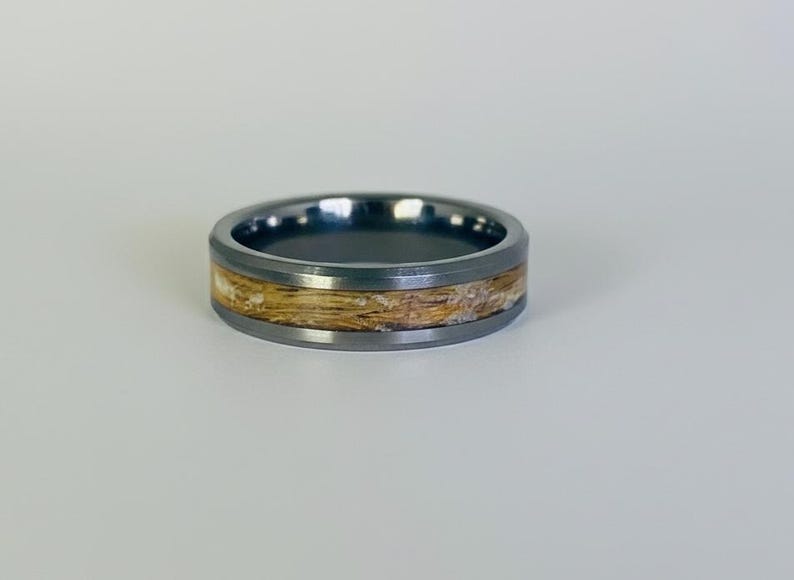 May include: A dark gray ring with a light-colored wood-like inlay. The ring has a smooth, polished finish and a simple, modern design. The band is approximately 8mm wide. This ring is suitable for everyday wear.