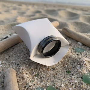 May include: A black ring with a silver stripe is displayed in a white paper container on a sandy beach. The ring is the focal point, with driftwood and sea glass scattered around. The background shows the ocean and sky.