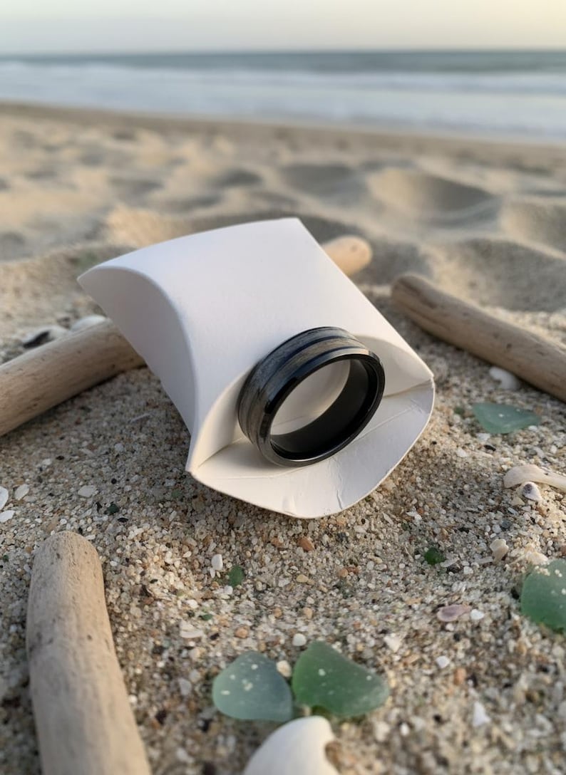 May include: A black ring with a silver stripe is displayed in a white paper container on a sandy beach. The ring is the focal point, with driftwood and sea glass scattered around. The background shows the ocean and sky.