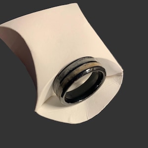 May include: A black ring with a band of gray and tan stripes, presented in a white paper holder. The ring has a polished finish and a modern design, suitable for everyday wear.