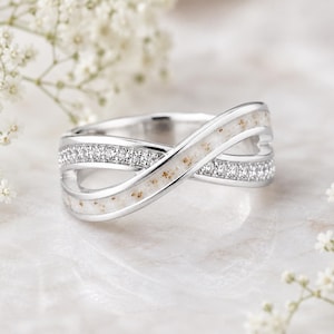 Könnte beinhalten: Ein silberfarbener Ring in Wickeloptik mit zwei ineinander verschlungenen Bändern. Ein Band ist mit kleinen, klaren Steinen besetzt, während das andere Band eine glatte, cremeweiße Einlage mit feinen beigen Sprenkeln auf der Oberfläche aufweist.