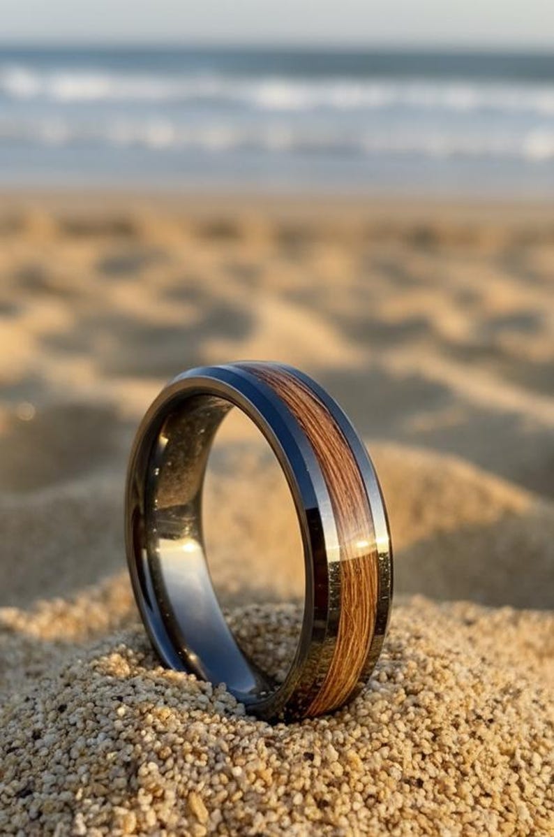 May include: A close-up of a ring with a dark gray band and a wood inlay. The ring is set on a sandy beach with the ocean in the background. The wood inlay is a light brown color.