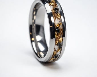 Può includere: Un anello in metallo color argento lucido con un canale centrale intarsiato riempito da un mix di fibre di capelli castani, piccoli frammenti di pietra bianca, pezzi di pietra nera e scaglie metalliche dorate.