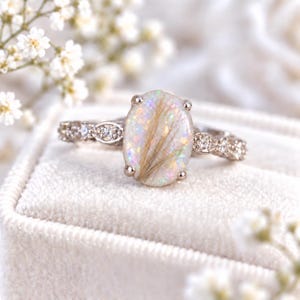 May include: A silver-toned ring featuring an oval-shaped center stone with an iridescent, pearlescent finish and visible golden-brown inclusions. The band is adorned with a series of small, clear, round-cut stones along the sides.