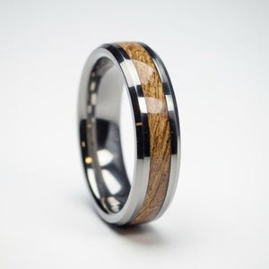 May include: A silver-colored ring with a wood inlay. The ring features a polished, metallic band with a central inlay of light brown wood. The wood grain is visible, adding texture and warmth to the design. A modern piece of jewelry.