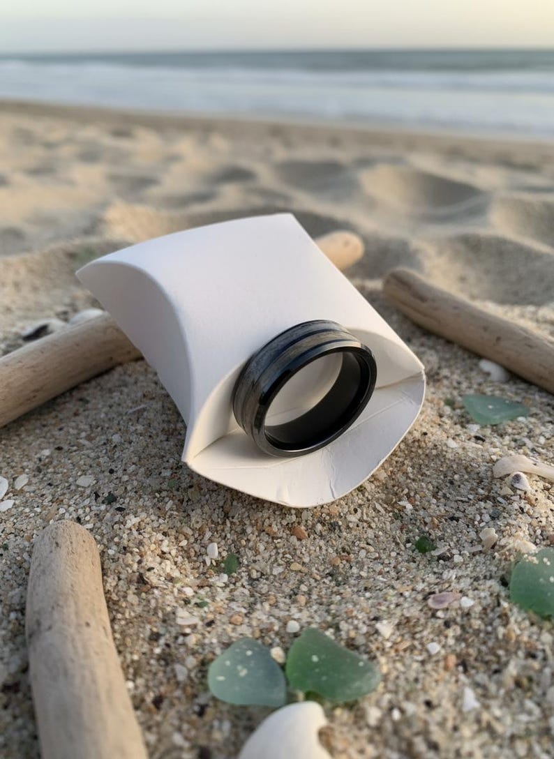 May include: A black ring with a silver stripe detail rests in a white paper container on a sandy beach. The ring is the focal point, with driftwood and sea glass scattered around. The ocean is visible in the background.