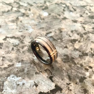 May include: A black and silver ring with a light wood grain and gold accents. The ring has a dark band and a light-colored center with two darker stripes and gold detailing.