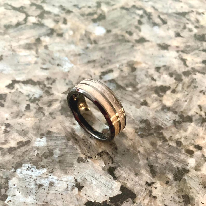 May include: A black and silver ring with a light wood grain and gold accents. The ring has a dark band and a light-colored center with two darker stripes and gold detailing.