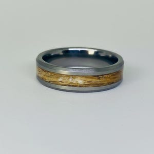 May include: A silver-toned ring with a band of light brown material. The ring has a dark gray outer band and a lighter gray inner band. The light brown material appears to be hair or a similar fibrous substance.