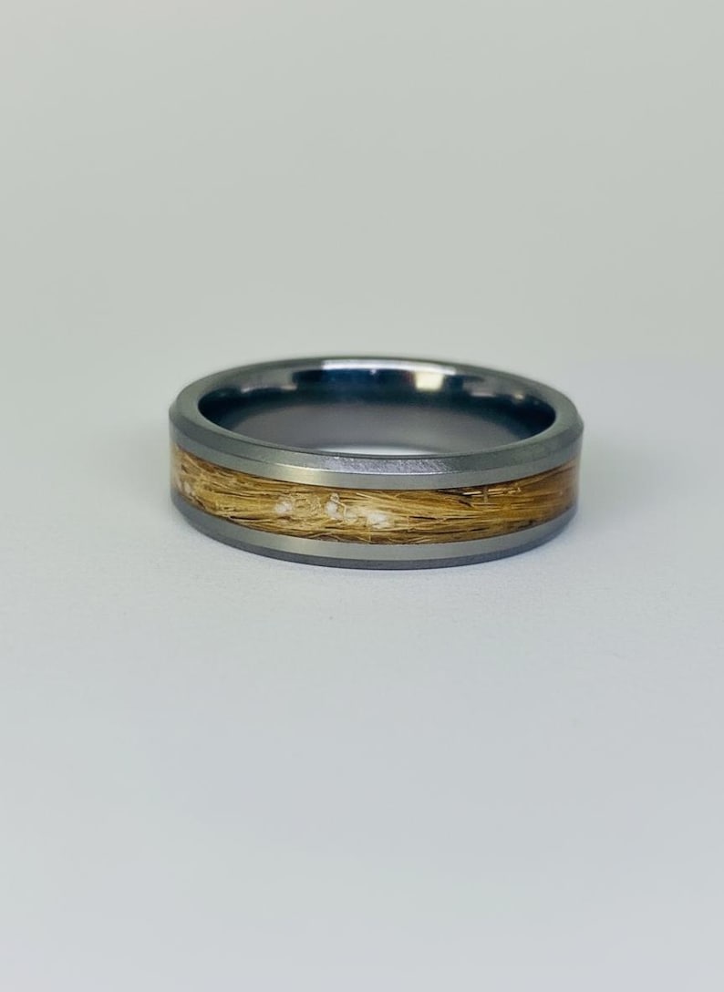 May include: A silver-toned ring with a band of light brown material. The ring has a dark gray outer band and a lighter gray inner band. The light brown material appears to be hair or a similar fibrous substance.