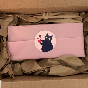 May include: A rectangular package wrapped in light pink paper, secured with a circular sticker featuring a black cat holding a red heart. The package is nestled inside a cardboard box lined with crumpled brown packing paper for protection.