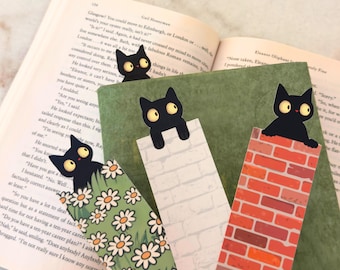 Peut inclure: Ensemble de marque-pages en forme de chat noir. Chaque chat noir est placé sur une surface différente : un mur de briques, un mur de briques blanches et un champ de marguerites. Les marque-pages sont dans un livre ouvert.