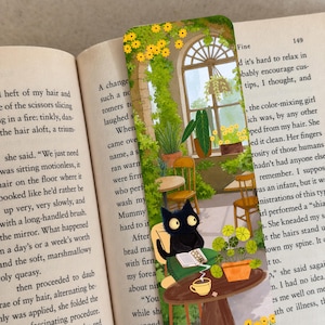 May include: A rectangular bookmark featuring a colorful illustration of a black cat sitting at a small round table in a sunlit greenhouse. The scene includes various potted green plants, yellow flowers, a steaming cup, and an open book on the table.