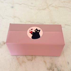 May include: A rectangular box wrapped in solid dusty pink paper, featuring a circular sticker on the center. The sticker displays a black cat with wide eyes holding a red heart, accompanied by two smaller red hearts on a light pink background.
