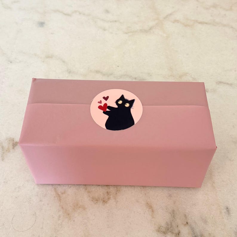 May include: A rectangular box wrapped in solid dusty pink paper, featuring a circular sticker on the center. The sticker displays a black cat with wide eyes holding a red heart, accompanied by two smaller red hearts on a light pink background.