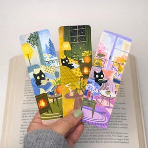 May include: Three rectangular bookmarks featuring illustrations of black cats reading books in cozy indoor settings. Each bookmark displays a different scene with warm lighting, furniture, plants, and windows, showcasing vibrant colors and detailed interior decor.