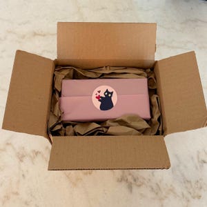 May include: An open cardboard shipping box containing a rectangular object wrapped in dusty pink paper. A circular sticker featuring a black cat holding a small red heart is centered on the wrapping paper, surrounded by crumpled brown packing paper.