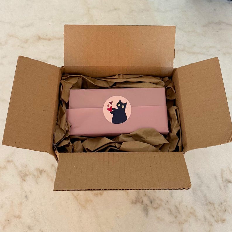 May include: An open cardboard shipping box containing a rectangular object wrapped in dusty pink paper. A circular sticker featuring a black cat holding a small red heart is centered on the wrapping paper, surrounded by crumpled brown packing paper.