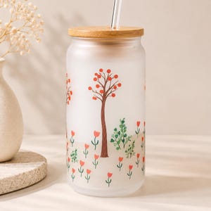 May include: A frosted glass tumbler with a bamboo lid and a clear glass straw. The exterior features a printed design of brown trees with orange circular leaves and small red tulip-like flowers scattered across the lower portion of the glass.