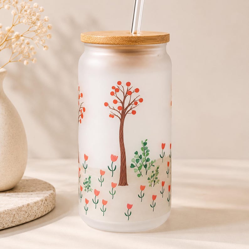 May include: A frosted glass tumbler with a bamboo lid and a clear glass straw. The exterior features a printed design of brown trees with orange circular leaves and small red tulip-like flowers scattered across the lower portion of the glass.