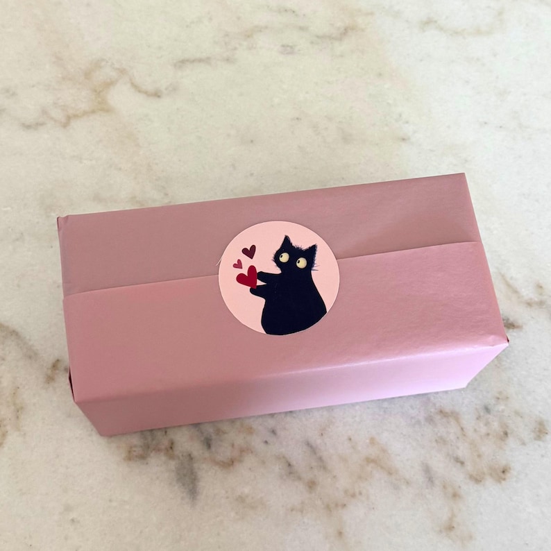 May include: A pink rectangular gift box wrapped with a matching band. A round sticker on the box features a black cat holding red hearts, with a pink background. The box is on a marble surface.