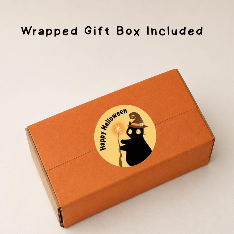 Puede incluir: Una caja de regalo rectangular de color naranja con una pegatina circular que muestra un gato negro con sombrero de bruja sosteniendo una varita brillante. La pegatina incluye el texto Happy Halloween. Arriba se lee Wrapped Gift Box Included.