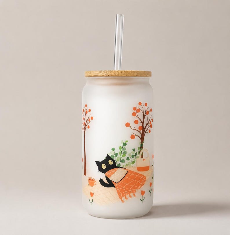 May include: A frosted glass tumbler with a bamboo lid and a clear glass straw. The exterior features a printed illustration of a black cat lying on a checkered blanket under orange-leafed trees, accompanied by a small bag and a cup.