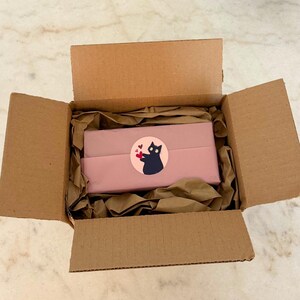 May include: Open cardboard box with pink wrapped packages and brown packing paper. A round sticker on the package features a black cat holding hearts. The box is on a light-colored surface.