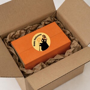 Puede incluir: Una caja rectangular naranja dentro de una caja de env&iacute;o de cart&oacute;n marr&oacute;n con papel marr&oacute;n arrugado. Una pegatina circular en la caja naranja muestra un gato negro con sombrero de bruja sosteniendo una varita, con el texto Happy Halloween.
