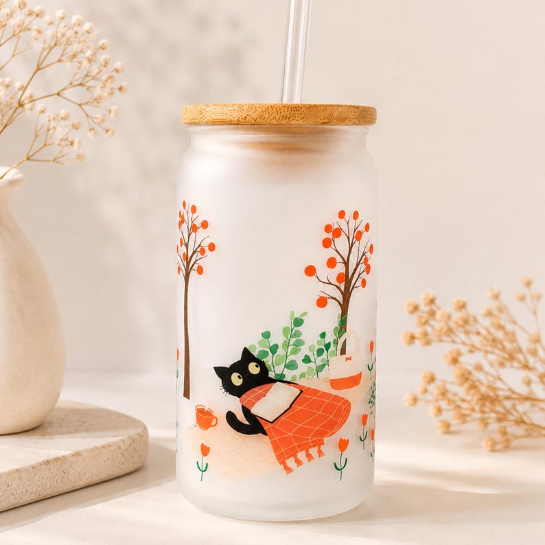 May include: A frosted glass tumbler with a bamboo lid and a clear straw. The exterior features a printed graphic of a black cat lying under an orange plaid blanket, surrounded by stylized trees with orange leaves, green bushes, and small orange flowers.