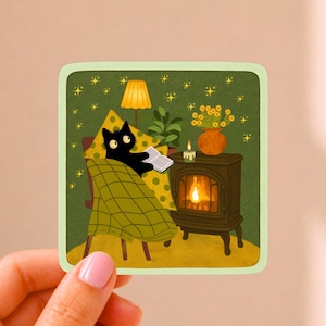 May include: A square sticker featuring a black cat sitting in a chair, covered by a green plaid blanket, reading an open book. Beside the cat is a dark brown wood stove with a glowing fire, a lit candle, a potted plant, and a yellow lamp.