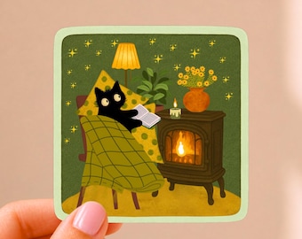 May include: A square sticker featuring a black cat sitting in a chair, covered by a green plaid blanket, reading an open book. Beside the cat is a dark brown wood stove with a glowing fire, a lit candle, a potted plant, and a yellow lamp.