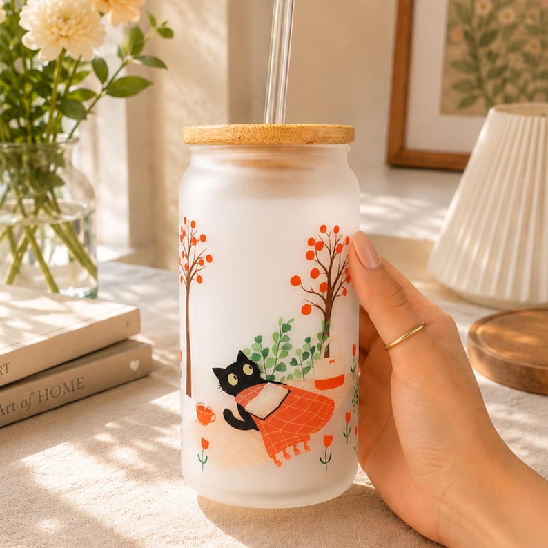 May include: A frosted glass tumbler with a bamboo lid and a clear glass straw. The exterior features a colorful graphic of a black cat lying under a blanket with a book, surrounded by stylized trees, small orange flowers, and a small bag.