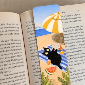 May include: A rectangular bookmark featuring a colorful illustration of a black cat lying on a blue and white striped beach towel. The scene includes a yellow and white beach umbrella, a small open book, slices of watermelon, and an orange soda can.