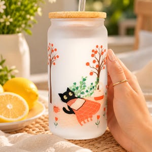 May include: A frosted glass tumbler with a bamboo lid and a clear straw, featuring a printed illustration of a black cat reading a book under orange trees, accompanied by a small cup and a tote bag on a light-colored checkered picnic blanket.