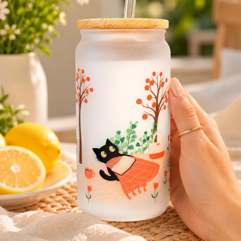 May include: A frosted glass tumbler with a bamboo lid and a clear straw, featuring a printed illustration of a black cat reading a book under orange trees, accompanied by a small cup and a tote bag on a light-colored checkered picnic blanket.