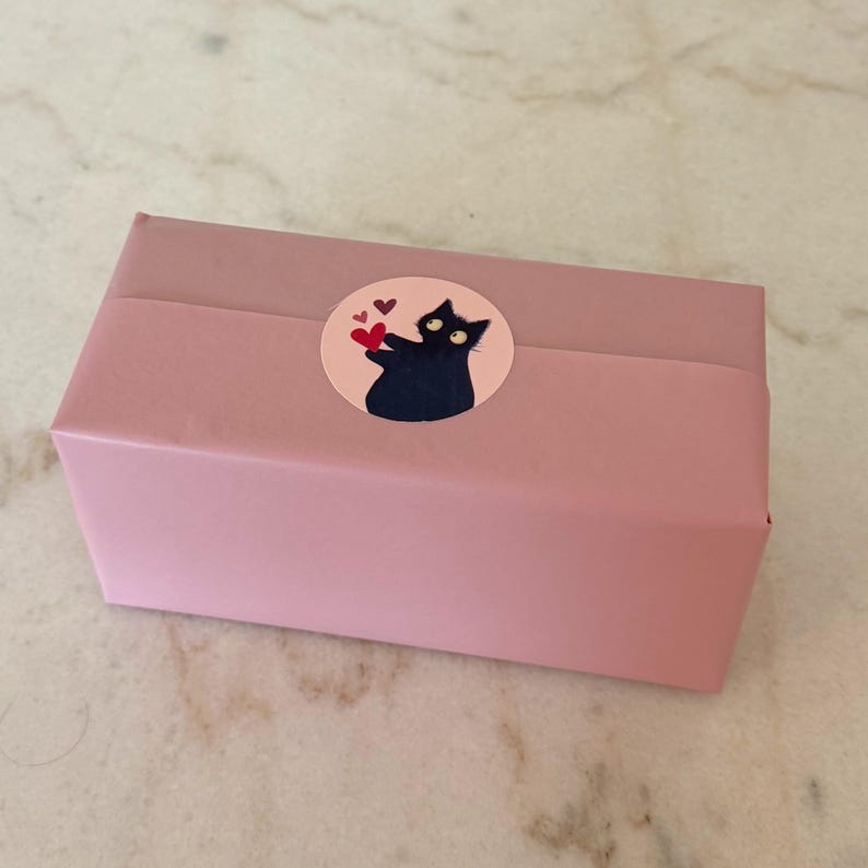 May include: A pink gift box wrapped with a matching band, adorned with a circular sticker. The sticker features a black cat holding hearts, with a few additional hearts around it. The box is on a marble surface.