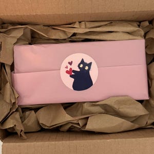 May include: A pink package wrapped in tissue paper, inside a cardboard box. A round sticker on the package features a black cat holding red hearts. The box is filled with brown packing paper.