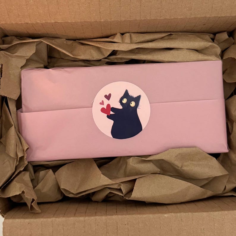 May include: A pink package wrapped in tissue paper, inside a cardboard box. A round sticker on the package features a black cat holding red hearts. The box is filled with brown packing paper.