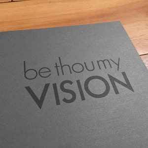 K&ouml;nnte beinhalten: Eine graue Karte mit den Worten "be thou my VISION" in dunklerem Grau gedruckt. Die Karte liegt auf einer Holzoberfl&auml;che.