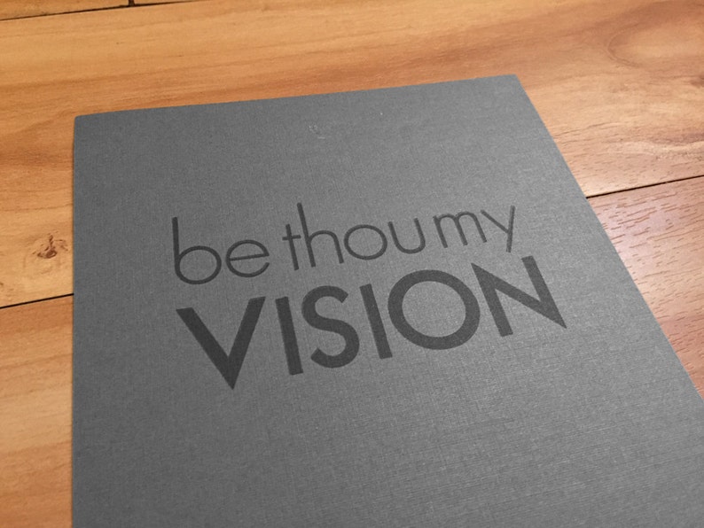 K&ouml;nnte beinhalten: Eine graue Karte mit den Worten "be thou my VISION" in dunklerem Grau gedruckt. Die Karte liegt auf einer Holzoberfl&auml;che.