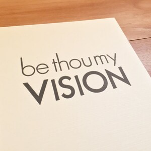 K&ouml;nnte beinhalten: Ein cremefarbener Druck mit den Worten "be thou my VISION" in dunkelgrauer, serifenloser Schrift. Der Druck befindet sich auf hellem Papier, vor einem Holzuntergrund.