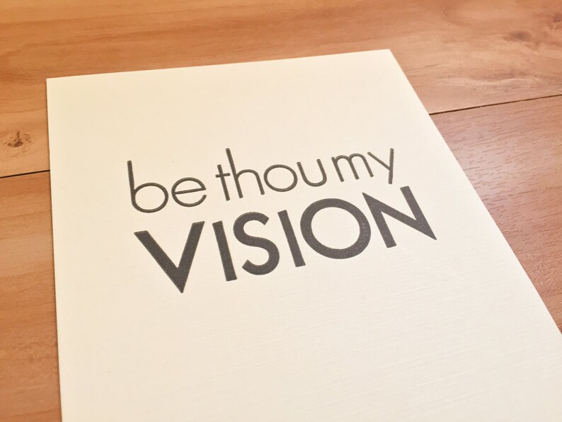 K&ouml;nnte beinhalten: Ein cremefarbener Druck mit den Worten "be thou my VISION" in dunkelgrauer, serifenloser Schrift. Der Druck befindet sich auf hellem Papier, vor einem Holzuntergrund.