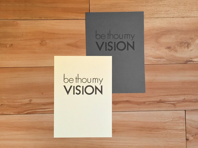 K&ouml;nnte beinhalten: Zwei rechteckige Karten mit dem Text "be thou my VISION" in fetter, schwarzer Schrift. Eine Karte ist cremefarben, die andere ist dunkelgrau. Die Karten liegen auf einer hellbraunen Holzoberfl&auml;che.