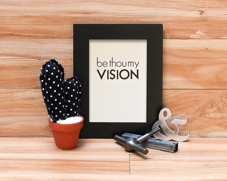 K&ouml;nnte beinhalten: Ein schwarzer gerahmter Druck mit den Worten "be thou my VISION" auf wei&szlig;em Hintergrund. Ein schwarz-wei&szlig; gepunkteter Kaktus in einem Terrakottatopf befindet sich links. Ein silbernes Et-Zeichen und ein Metallwerkzeug befinden sich rechts.