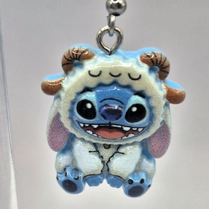 Puede incluir: Un pendiente azul y blanco con el tema de Stitch. El pendiente presenta un personaje de Stitch sonriente que lleva una capucha peluda con cuernos marrones. El pendiente est&aacute; sujeto a un anzuelo plateado.