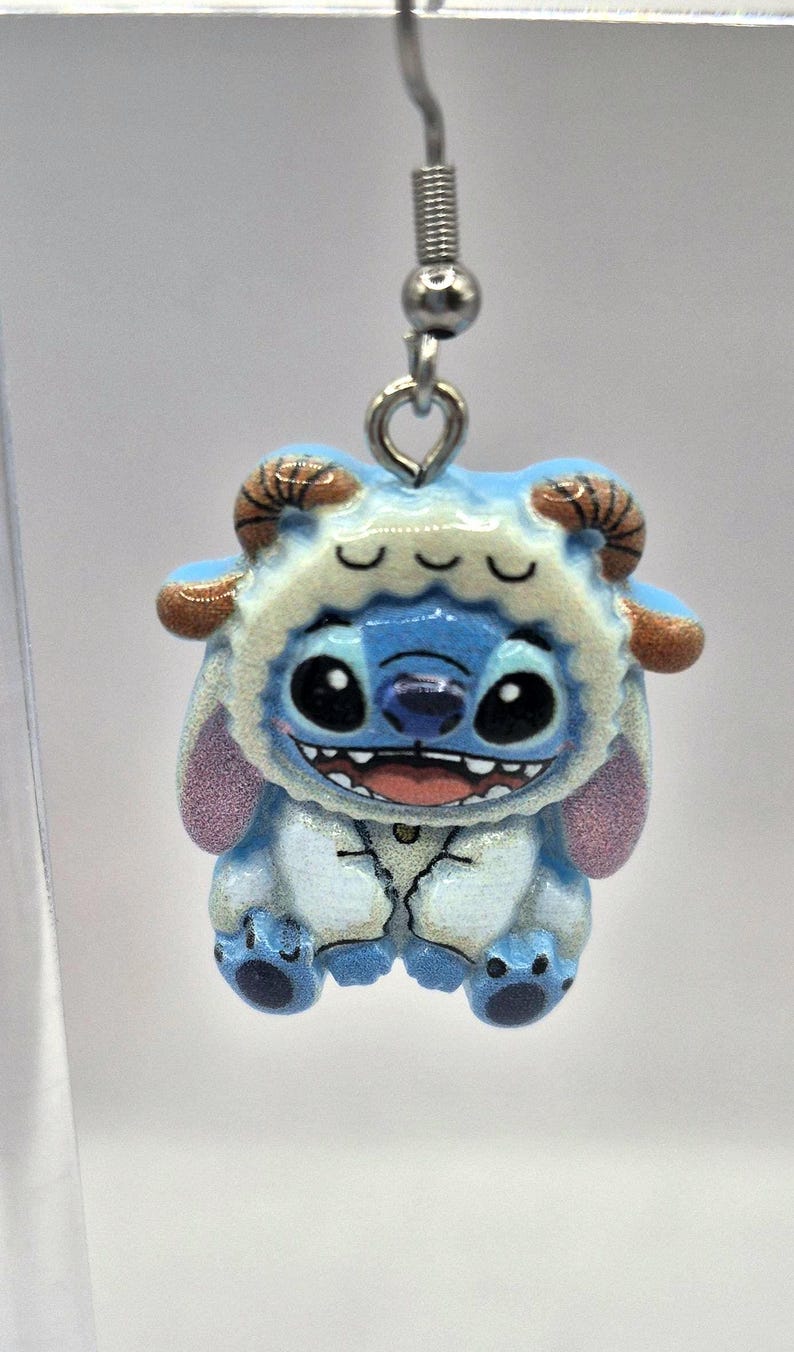 Puede incluir: Un pendiente azul y blanco con el tema de Stitch. El pendiente presenta un personaje de Stitch sonriente que lleva una capucha peluda con cuernos marrones. El pendiente est&aacute; sujeto a un anzuelo plateado.