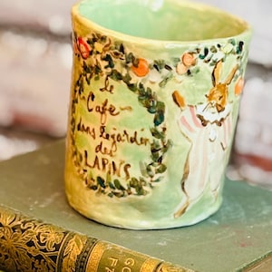 May include: A light green ceramic mug with a hand-painted illustration of a rabbit wearing a striped outfit. The mug features a floral wreath surrounding the text "le Cafe dans le jardin des LAPINS" in brown script, with orange accents.